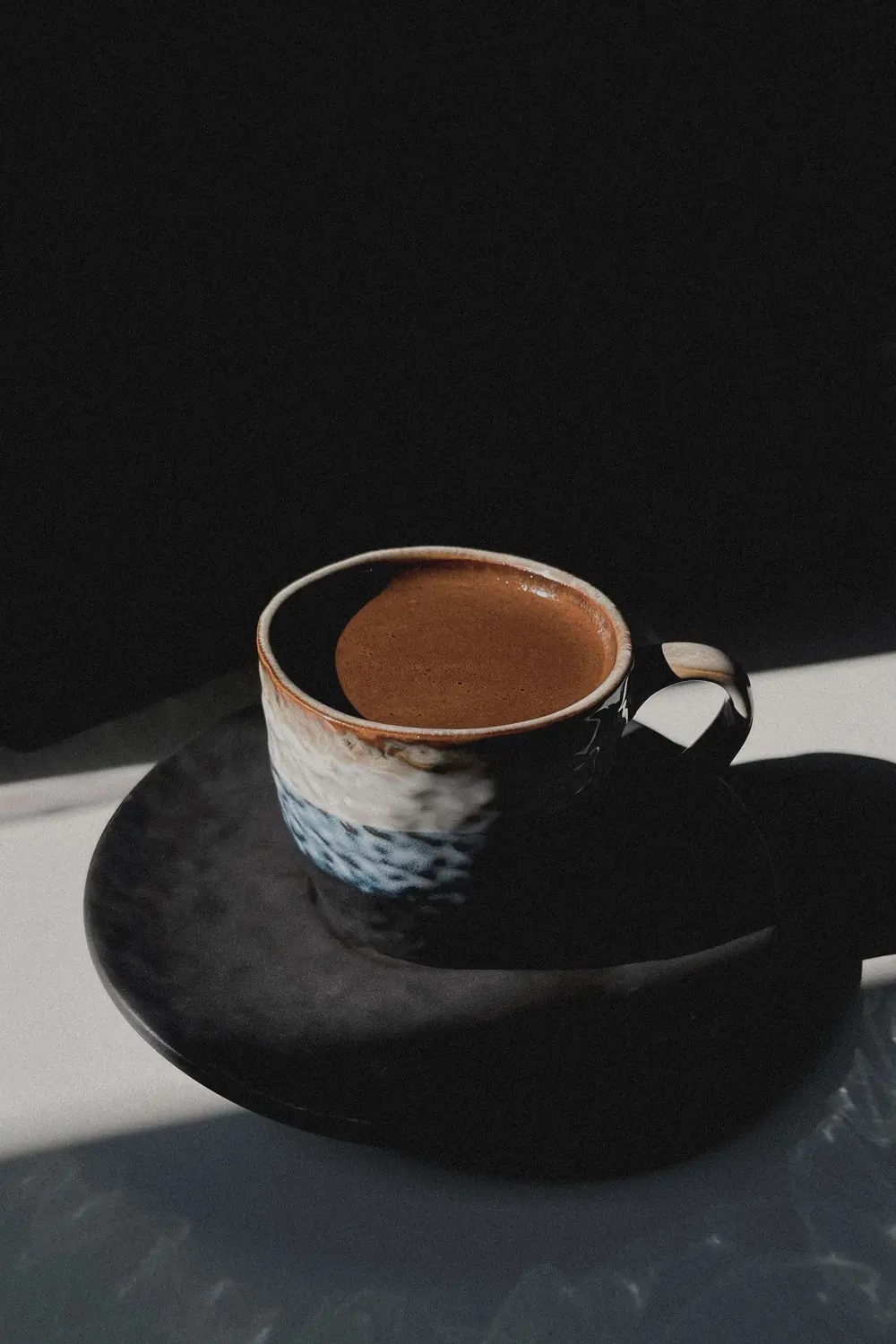 A glazed stoneware coffee cup on a dark saucer, caught in a single shaft of light