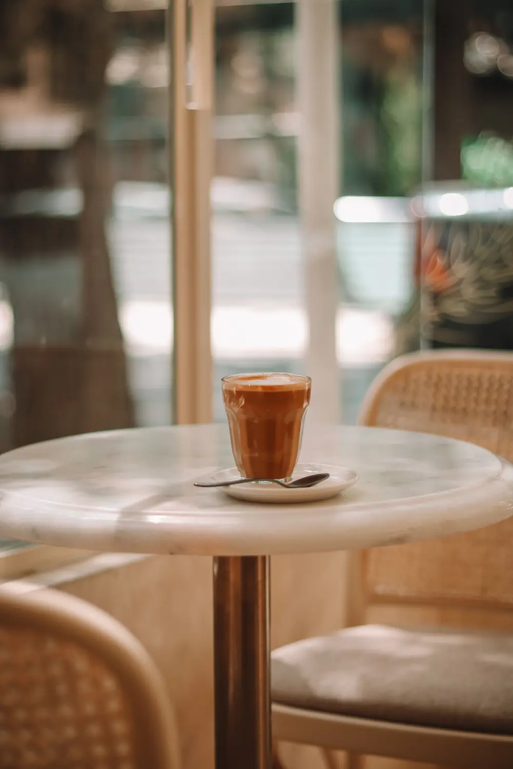 A cappuccino on a small marble café table by a sunny window, rattan chairs behind